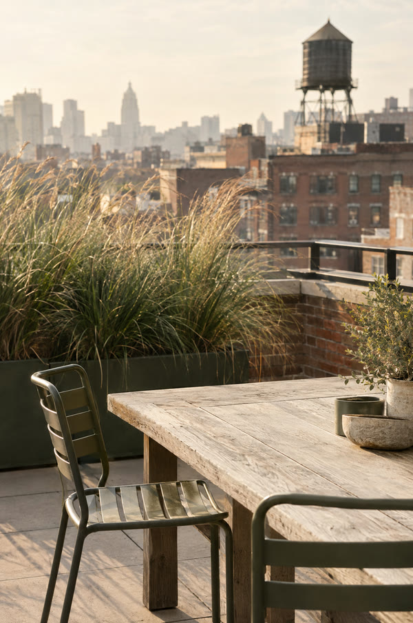 A rooftop terrace in late afternoon with an oak table, metal chairs and a planter, city facades soft in the background. Illustrative image.